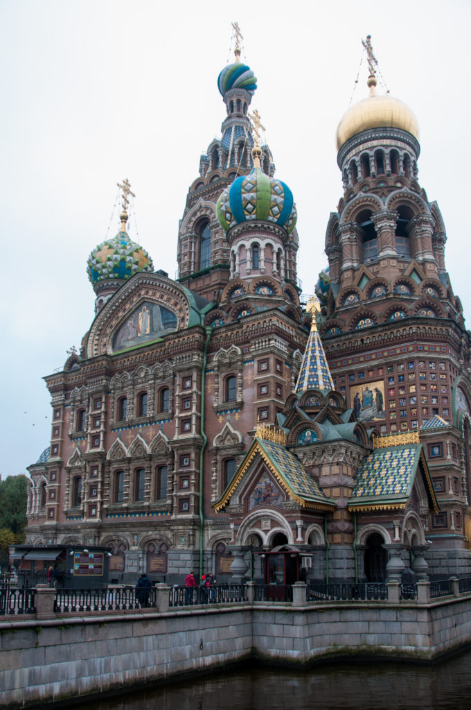 Oblique canal-side view of the church with clustered brick towers, colorful onion domes, mosaic gables, patterned roofs, and pedestrians.
