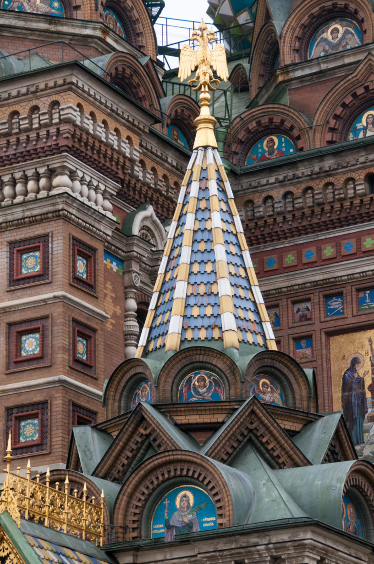 Blue-and-cream tiled pyramidal roof with gilded finial surrounded by brick arches, mosaics, carved bands, and gold cresting.
