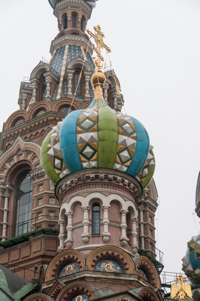 Turquoise, green, and ocher onion dome with geometric panels above an arcaded drum, backed by brick towers and crosses.