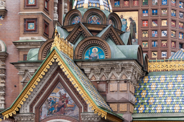 Gilded triangular gable with religious mosaic amid brick arches, mosaic panels, carved masonry, and multicolored roof tiles.