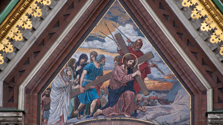Triangular exterior mosaic showing Christ carrying the cross amid a group of figures, framed by dark brick and gilded ornament.