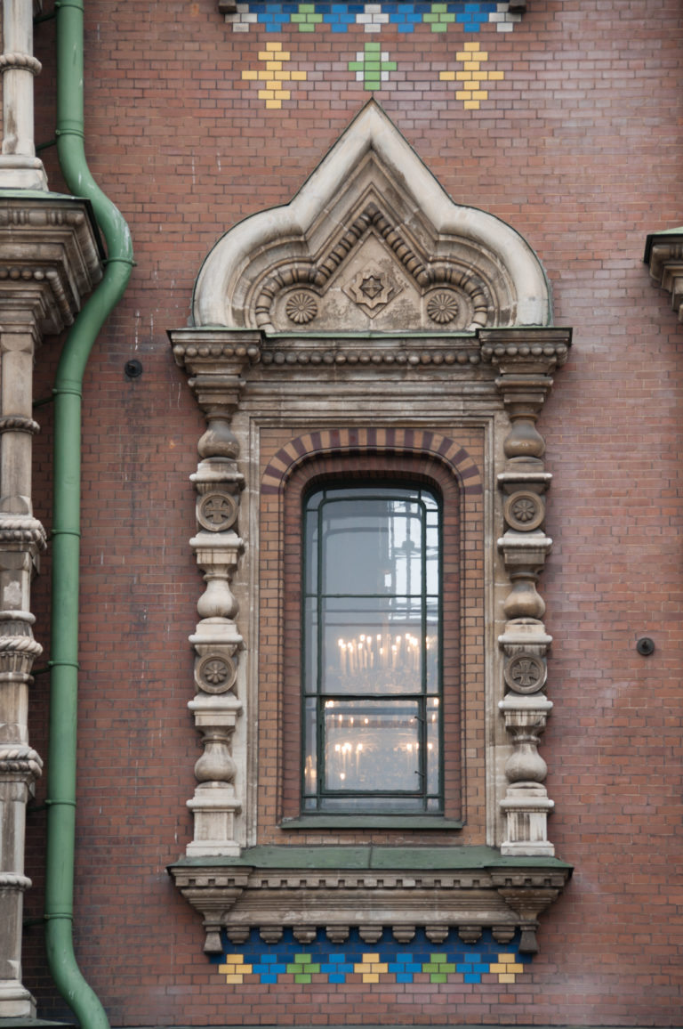 Rectangular window with ornate pale stone surround in a red-brick wall, beside a green downpipe and colored tile bands.