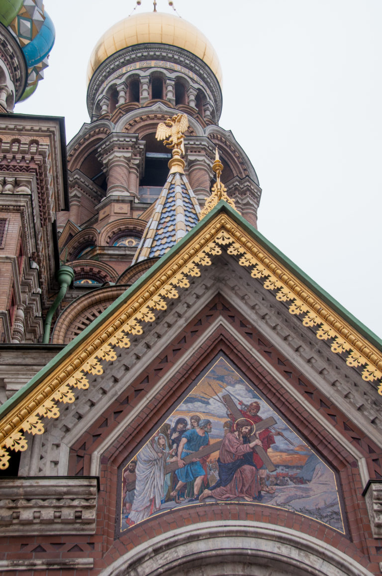 Triangular religious mosaic beneath gilded roof trim, with patterned brickwork and a domed cylindrical tower rising behind.