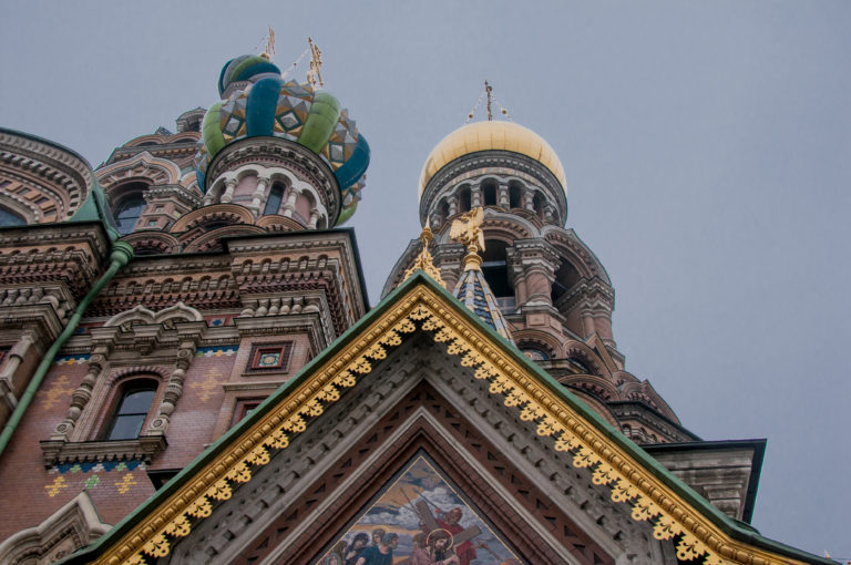 Low-angle exterior detail of a mosaic-filled triangular gable beneath colorful onion domes and a cylindrical church tower.