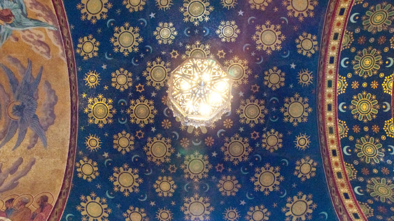 Deep blue vaulted ceiling patterned with gold stars and sunbursts around a bright chandelier, with figurative mosaics at the edges.