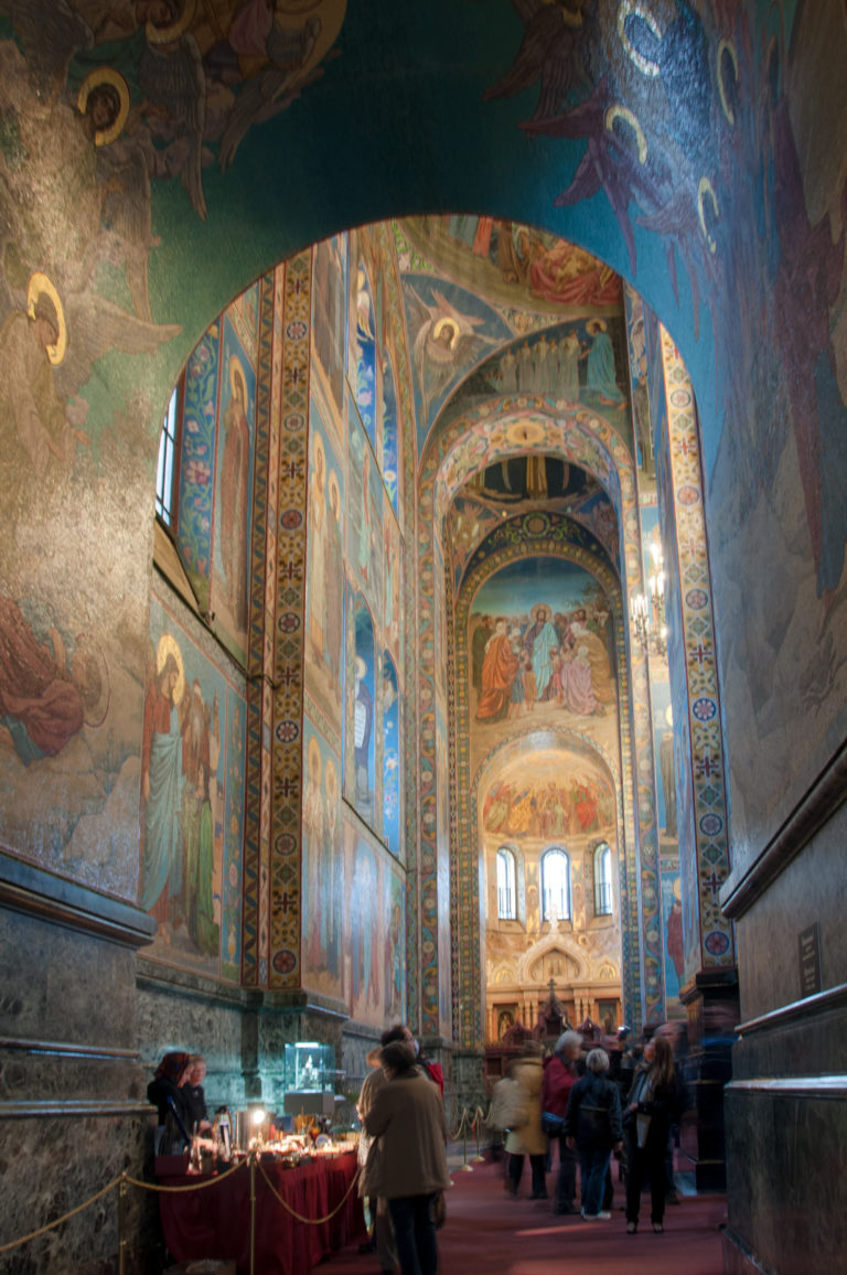 Long view through mosaic-covered arches and piers inside the church, with visitors and a small table in the foreground.
