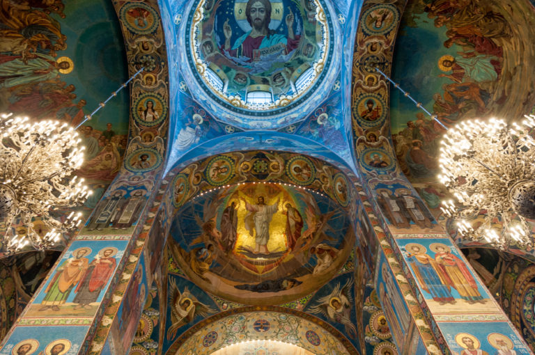 Steep centered view of mosaic-covered vaults with Pantocrator above, Transfiguration below, and bright chandeliers on both sides.