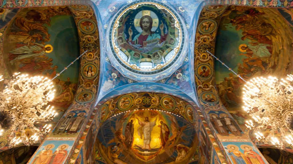 Centered upward view linking Christ Pantocrator above with a radiant Transfiguration mosaic below, flanked by chandeliers.