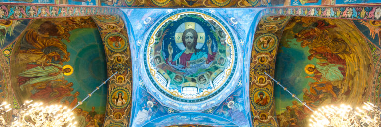Oblique upward view of the Pantocrator mosaic, adjacent figurative vaults, and bright chandeliers inside the church.