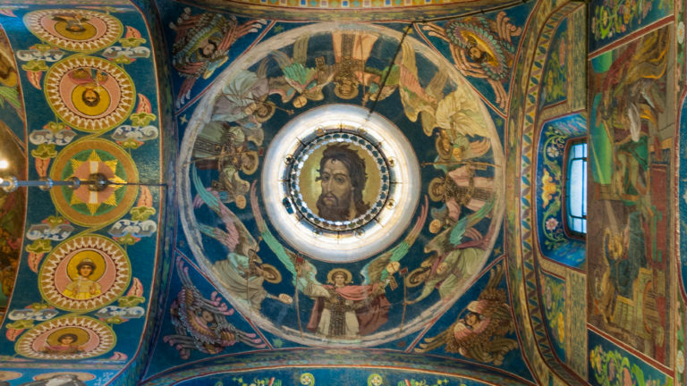 Upward view of a circular Christ mosaic centered in an angel-filled vault, with ornamental medallions and a window at right.