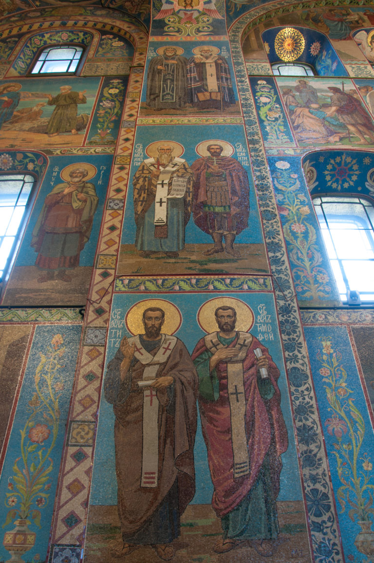 Full-length haloed saints in stacked wall mosaics, including two vested clerical figures between patterned borders and windows.