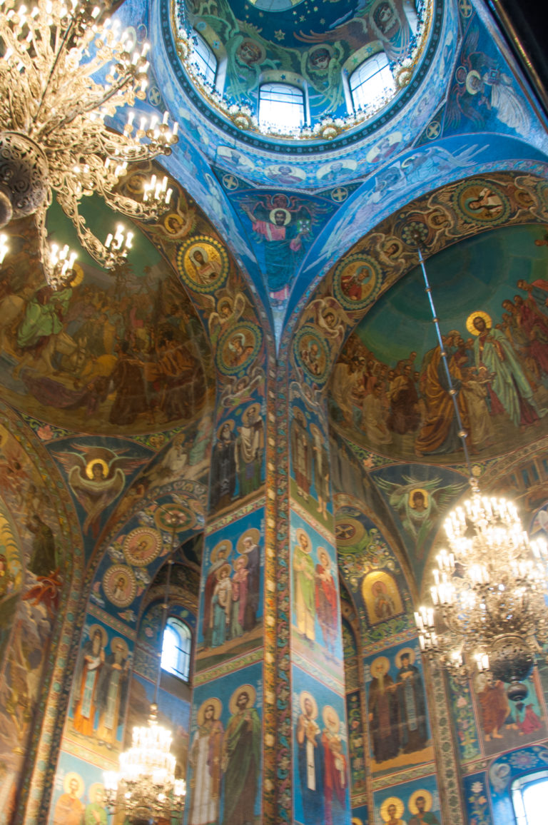 Angled interior view of mosaic-covered piers, arches, and vaults with bright chandeliers hanging between the decorated bays.
