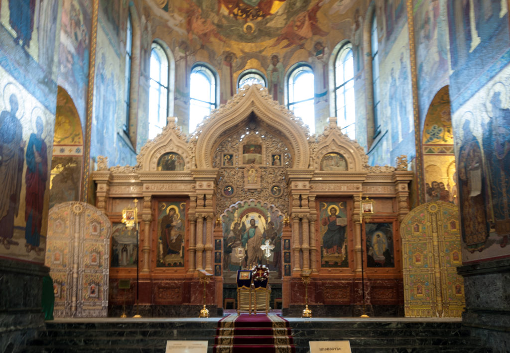 Frontal view of the three-gabled iconostasis in a tall mosaic-lined apse with windows and dark stone lower walls.