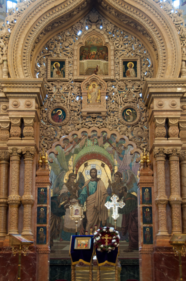 Ornate pale-stone iconostasis with carved arches and icons before a large mosaic of Christ in the sanctuary.