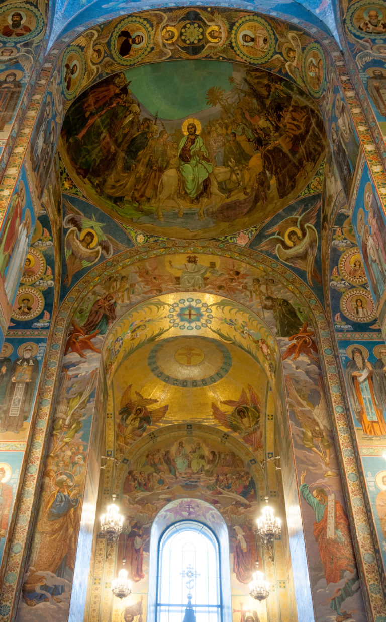 Upward view of a curved mosaic-covered vault above a bright window, with narrative scenes and circular saint medallions.