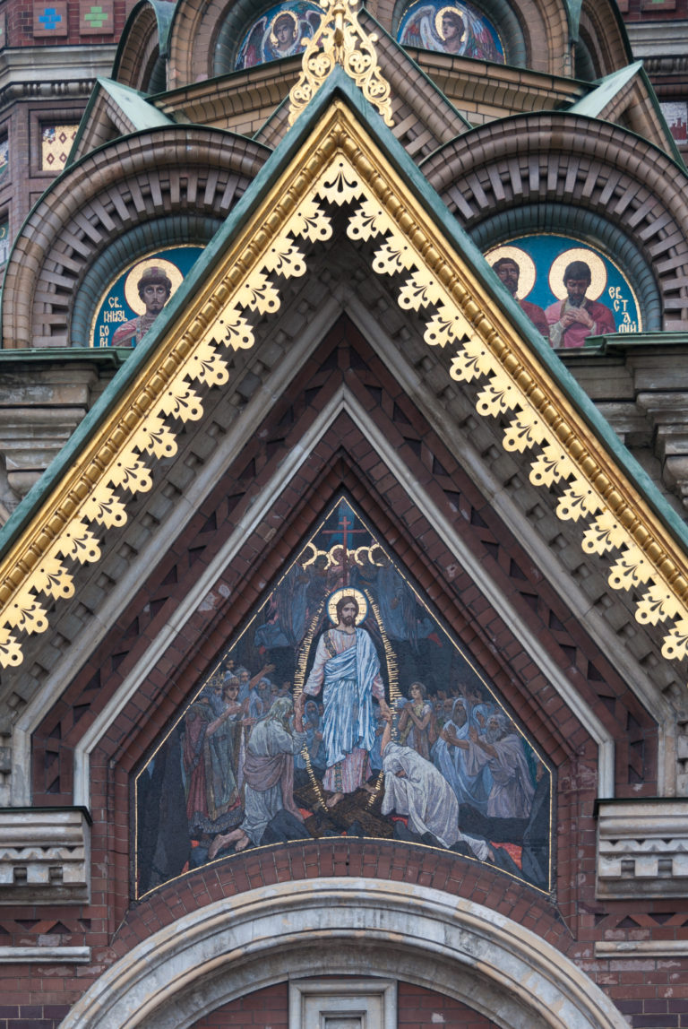 Triangular gable on the church exterior containing a religious mosaic, with patterned brickwork, gilded trim, and saint medallions above.