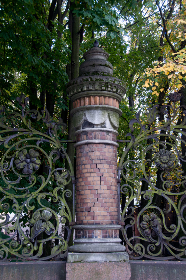 Red-brick gate pier with stone bands beside ornate wrought-iron fencing and leafy trees at the church grounds.