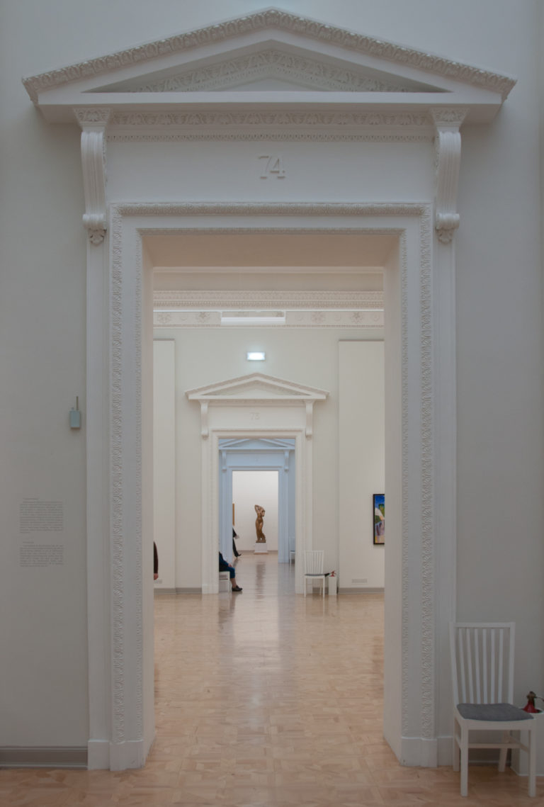 White pedimented doorways aligned through several museum galleries, with parquet floors and a small sculpture in the distance.