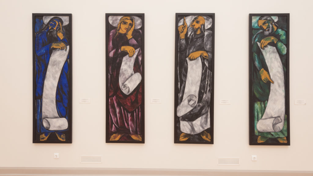 Four narrow vertical paintings showing elongated robed figures in blue, purple, ocher, white, black, and green.