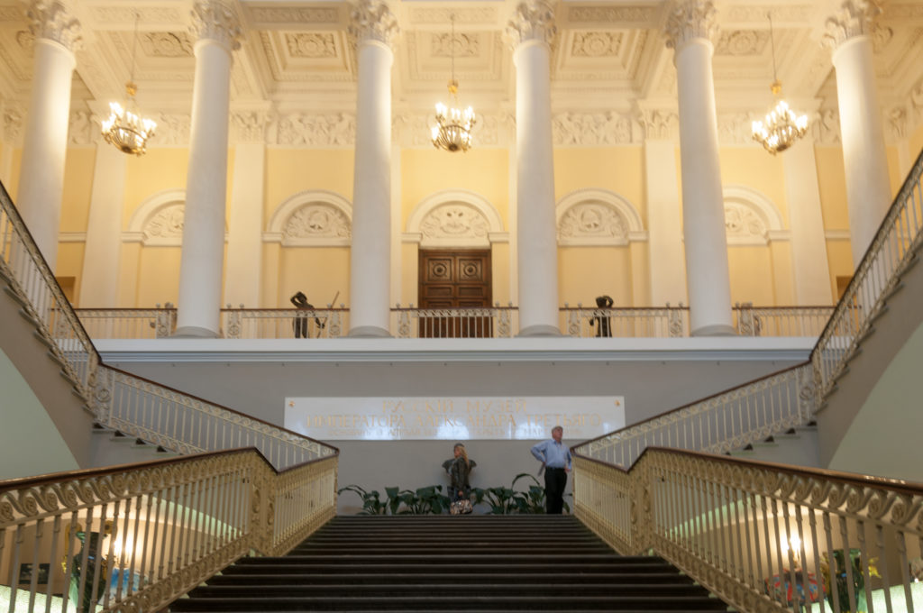 Broad staircase with gilded rails leading to white columns and a central doorway, with visitors near the landing.