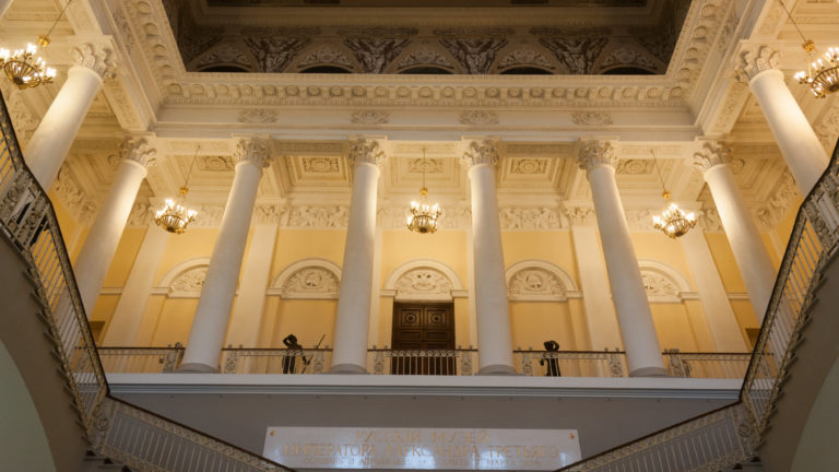 Upward view of tall white columns, chandeliers, and ornate ceiling above the museum staircase.