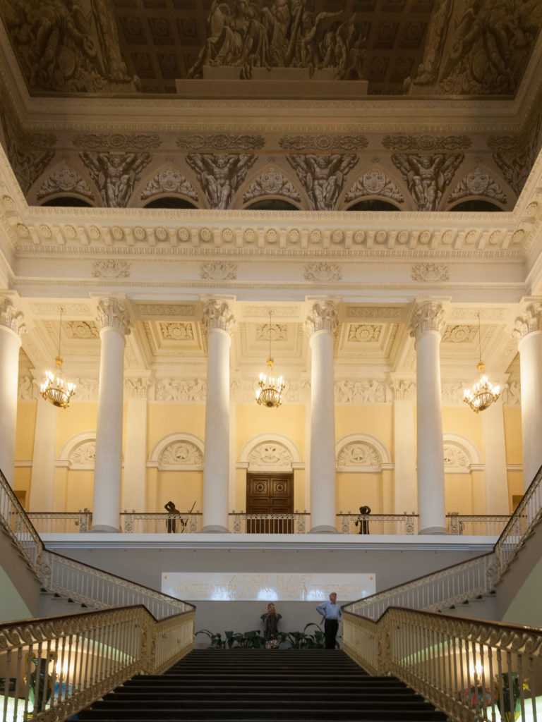 Symmetrical grand staircase with gilded railings rising to a landing lined with tall white columns.