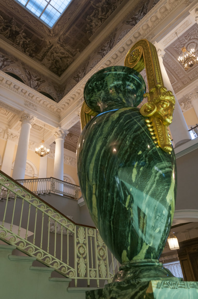 Large polished green stone urn with gilded mounts beside an ornate staircase and white columns.