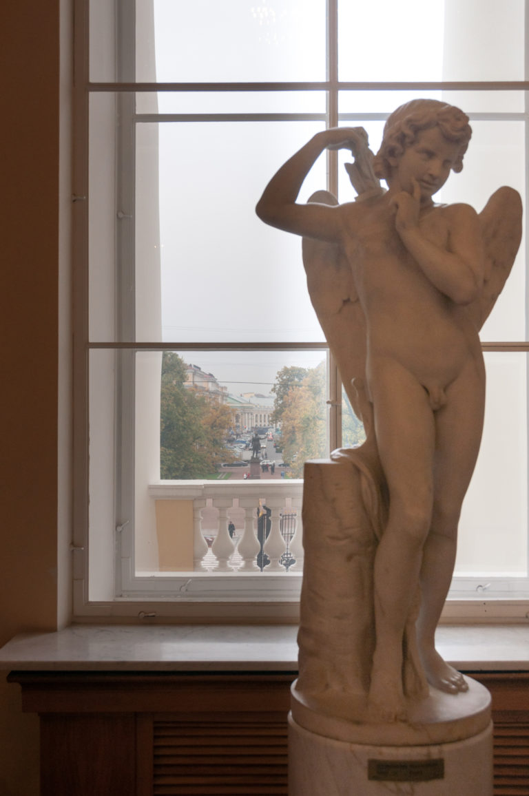 Winged child sculpture playing a transverse flute in front of a tall museum window.