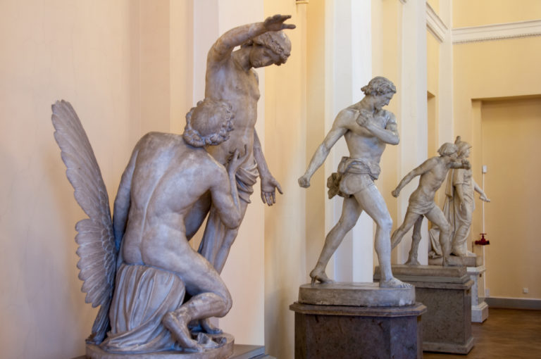 Row of classical-style sculptures on pedestals, led by a large kneeling winged figure in the foreground.