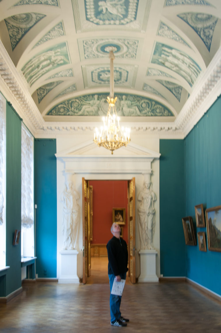 Visitor standing in a blue museum gallery facing a white pedimented doorway leading to a red room.
