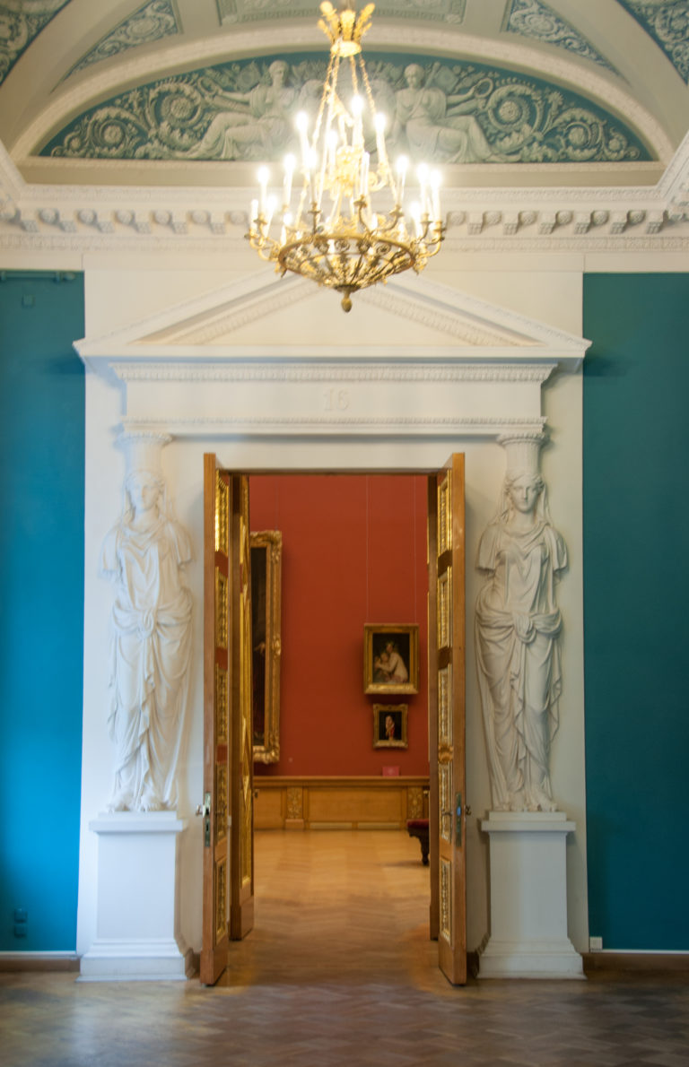 Pedimented doorway with flanking white statues between a blue gallery and a red gallery beyond.