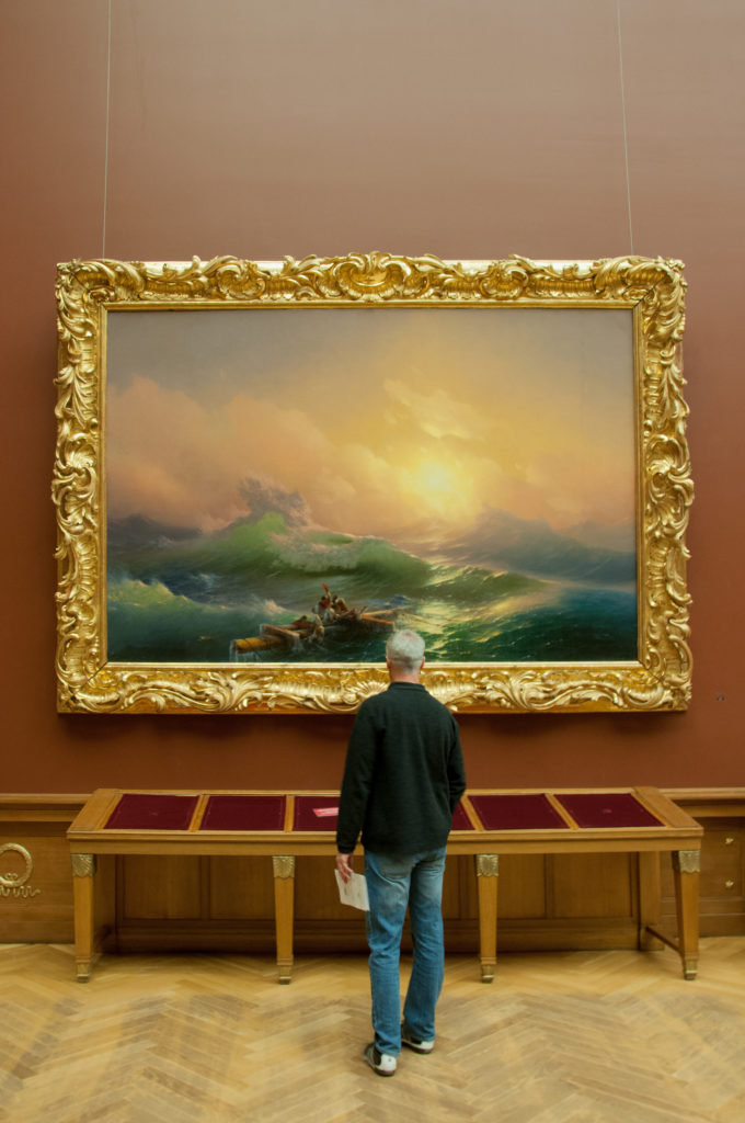 Visitor viewing Ivan Aivazovsky’s 1850 The Ninth Wave, where shipwreck survivors cling to wreckage amid huge green waves and a glowing sunrise.