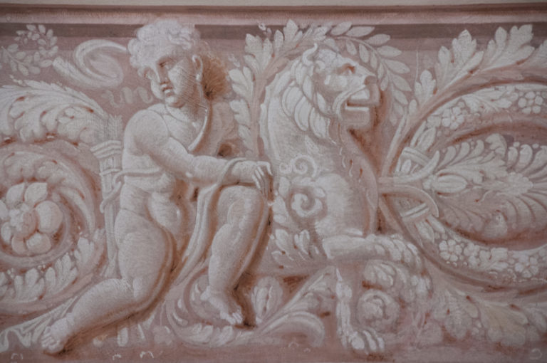 Pale decorative relief of a seated classical figure surrounded by scrolling leaves, flowers, and symmetrical ornamental motifs.