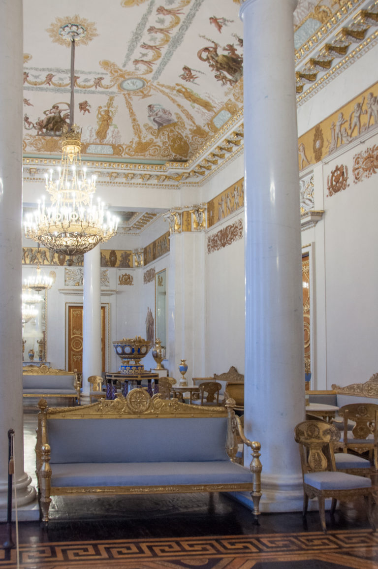 Long ornate palace salon with upholstered sofas, white columns, chandeliers, painted ceiling, gilded trim, and parquet floor.