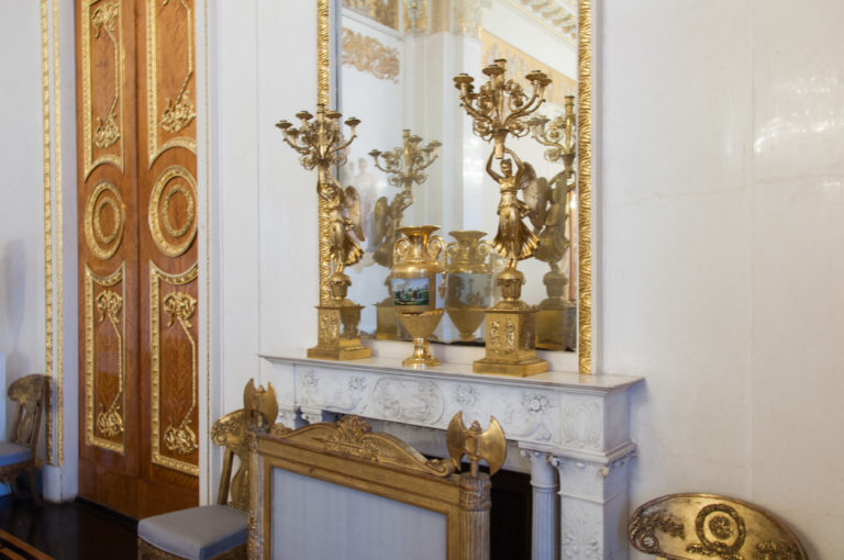 White marble mantel with gilded candelabra and vessels beside richly decorated double doors and a tall mirror.