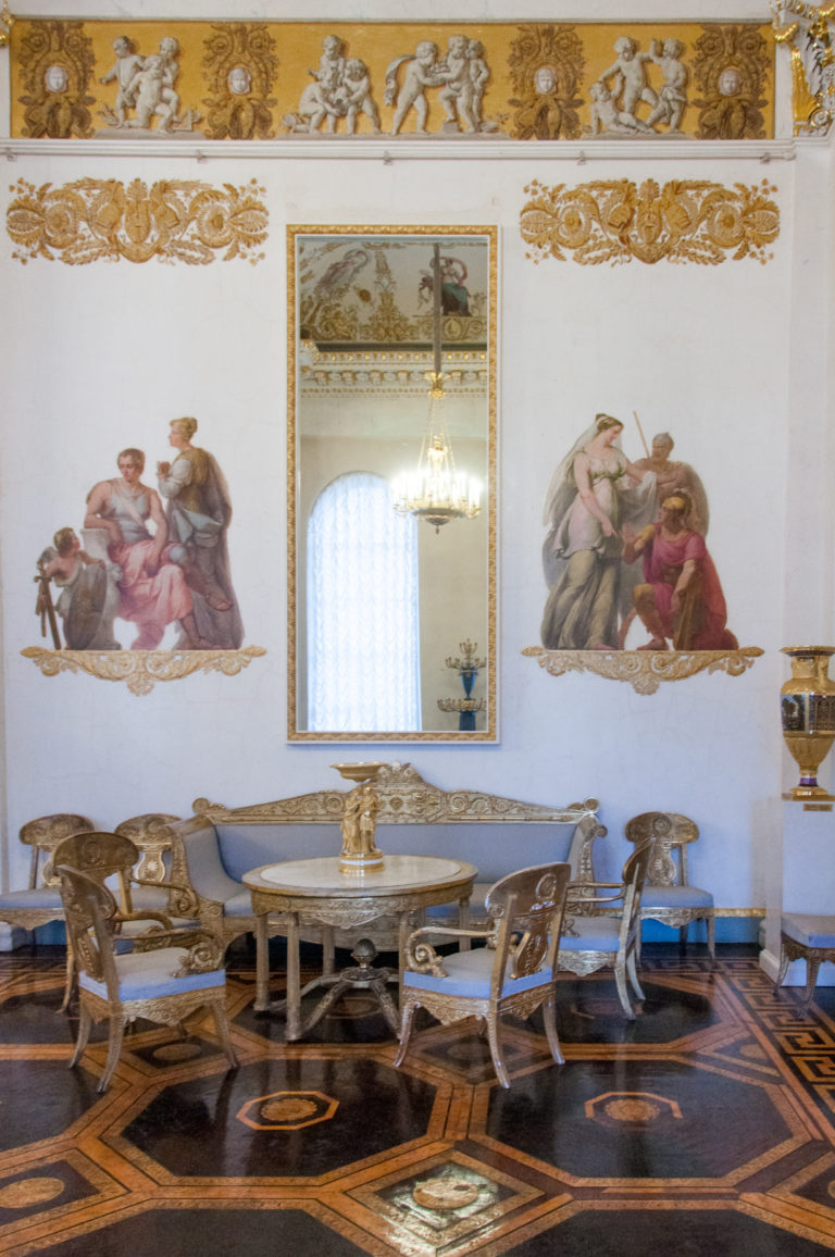 Round table and carved chairs in an ornate palace salon with wall paintings, gilded frieze, mirror, chandelier, and parquet floor.