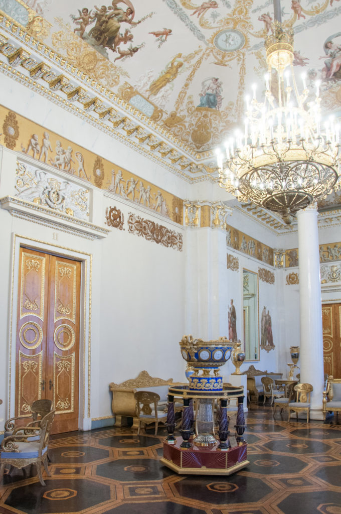 Ornate palace salon with painted ceiling, chandelier, white columns, parquet floor, carved furniture, and a blue-and-gold vessel.
