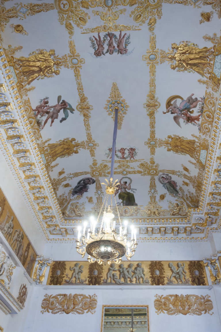 Painted mythological figures and gilded ornament cover a rectangular palace ceiling above a glowing crystal chandelier.