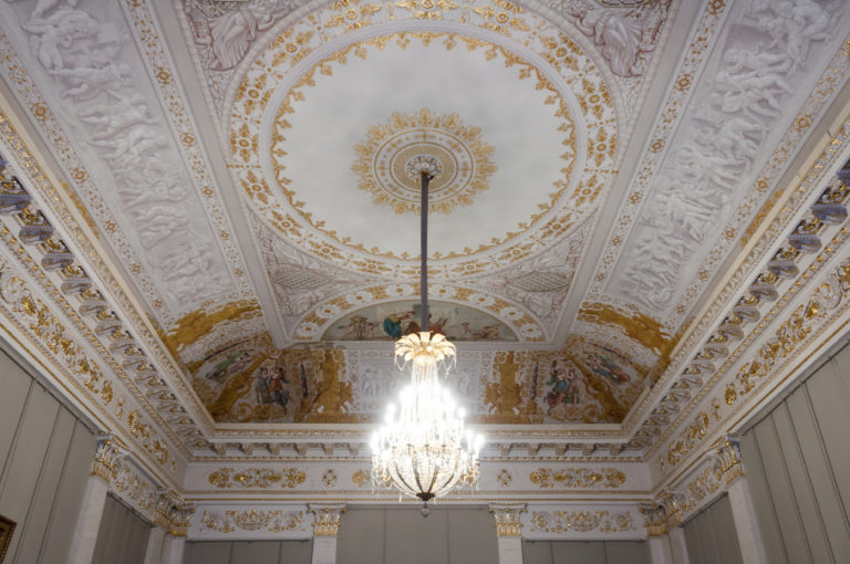 Crystal chandelier hangs beneath an ornate white-and-gold ceiling with a circular rosette and painted figural frieze.