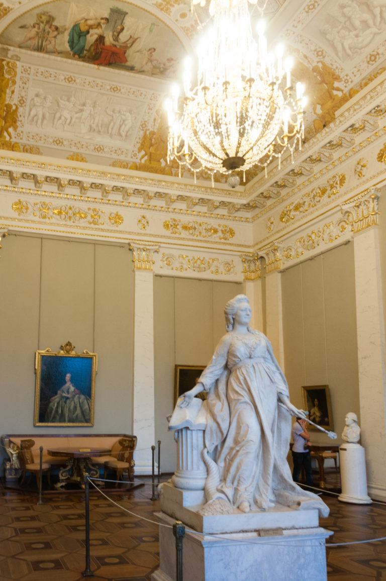 White marble seated figure seen from behind in an ornate gallery with chandeliers, gilded ceiling decoration, and framed paintings.