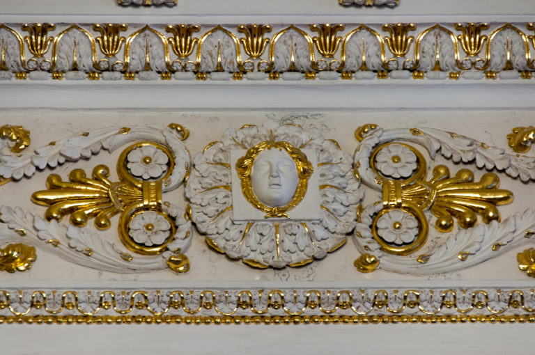 White-and-gold stucco frieze with a female mask, rosettes, scrolling leaves, and beaded borders in a palace interior.