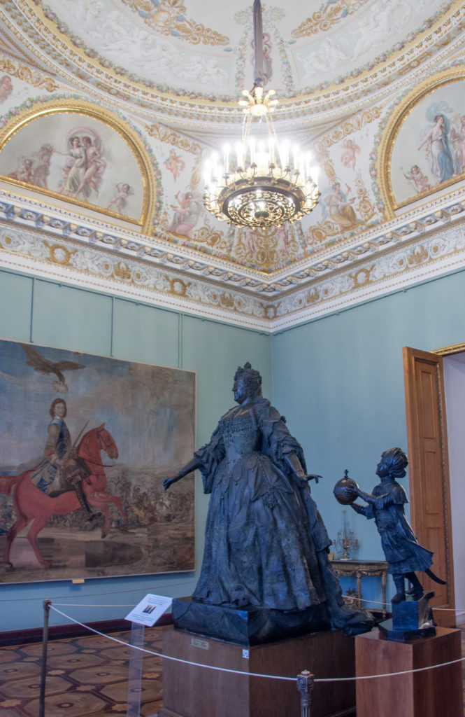 Ornate palace gallery with a large dark bronze sculpture, painted wall panels, chandelier, gilded ceiling decoration, and parquet floor.