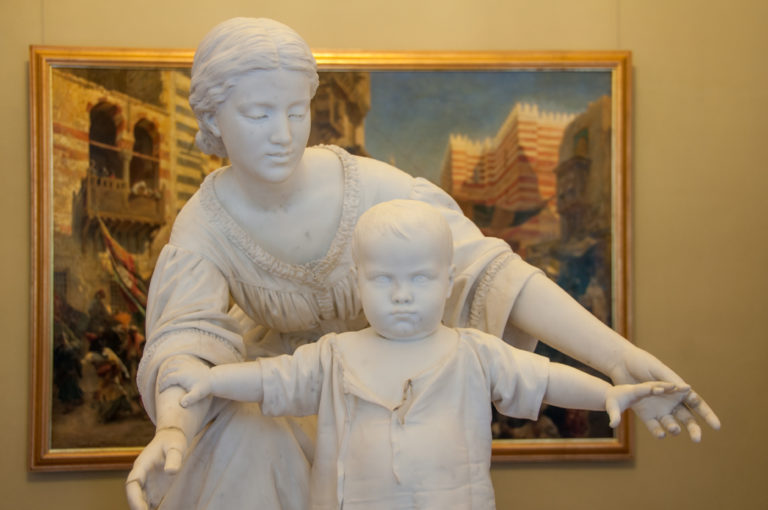 White marble sculpture of a woman supporting a small child who stands in front with both arms stretched outward.