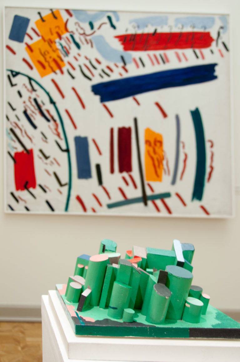 Abstract painting above a green geometric sculpture composed of clustered cylindrical and block forms in a museum gallery.