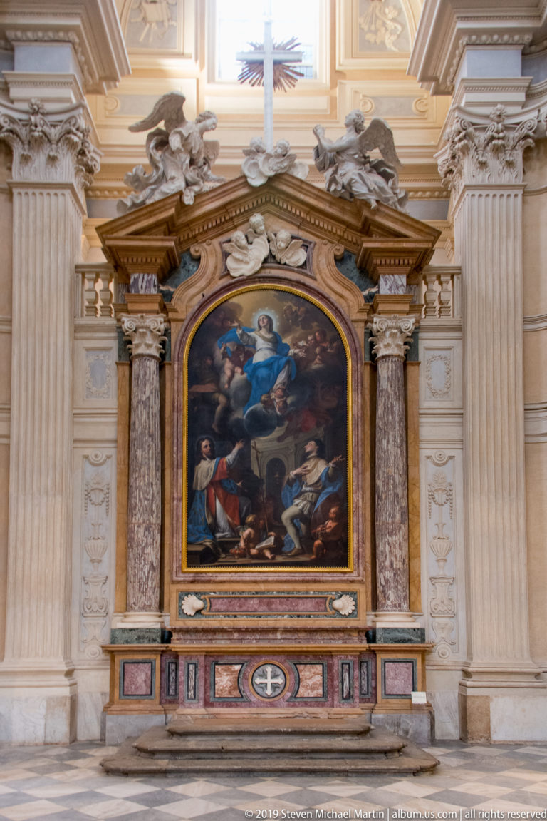 Ornate chapel altar with colored marble, gilded trim, sculpted angels, and a large religious painting in a pale architectural surround.