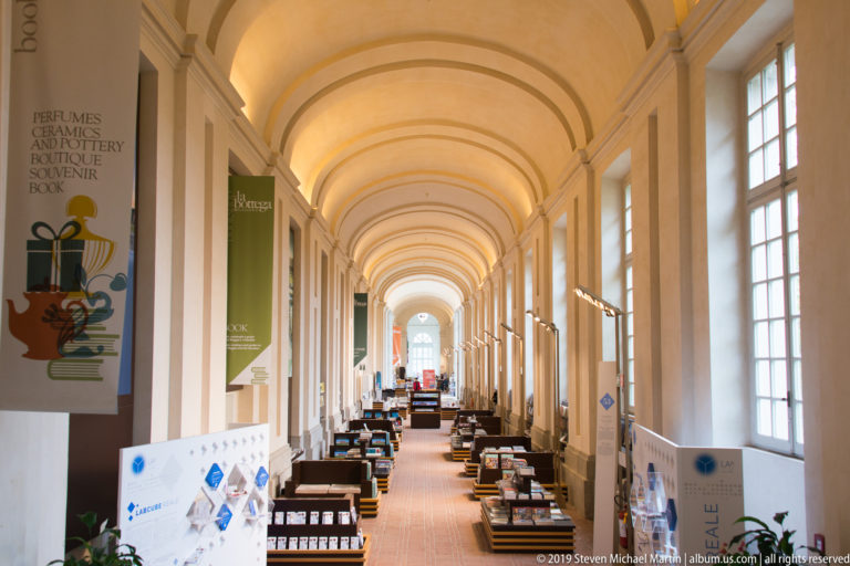 Long vaulted palace corridor with tall windows, exhibition tables, banners, and display cases arranged along a central aisle.