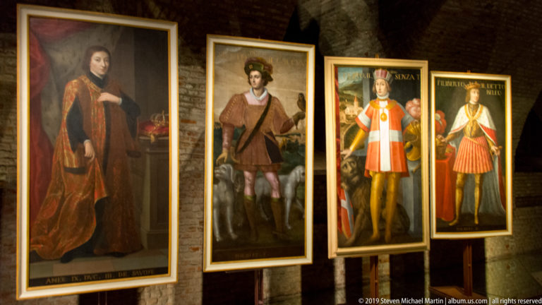 Four tall historical portrait paintings displayed side by side in a brick-vaulted gallery at La Venaria Reale.
