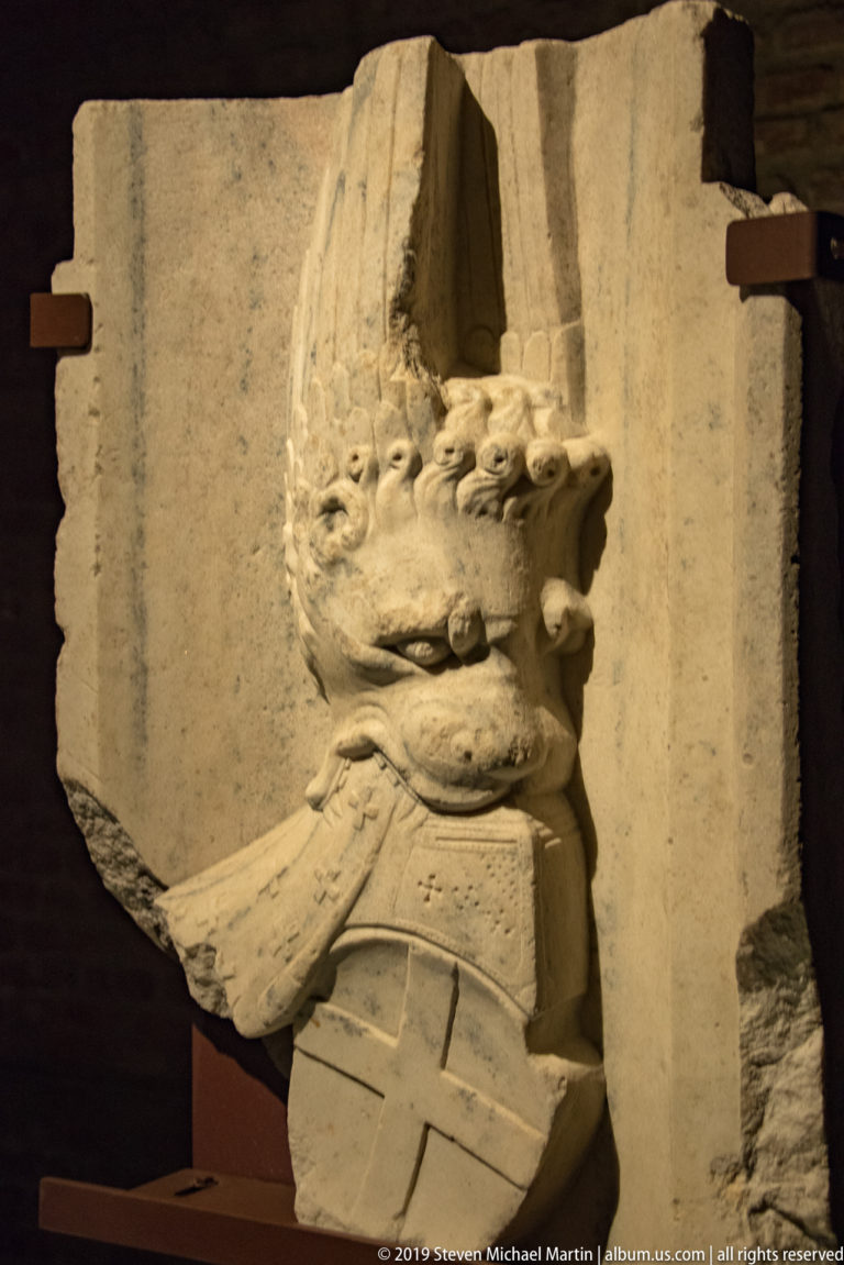 Weathered stone relief fragment with a profile head in a tall crested helmet, mounted against a dark museum background.