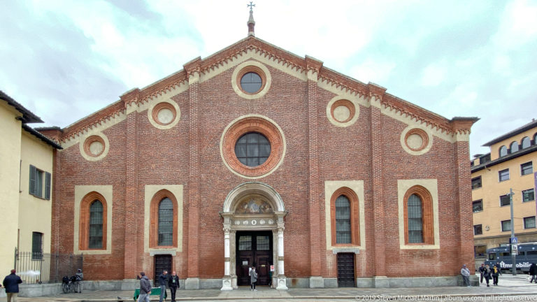Full frontal view of Santa Maria delle Grazie with red-brick gable, circular windows, stone portal, and visitors in the piazza.