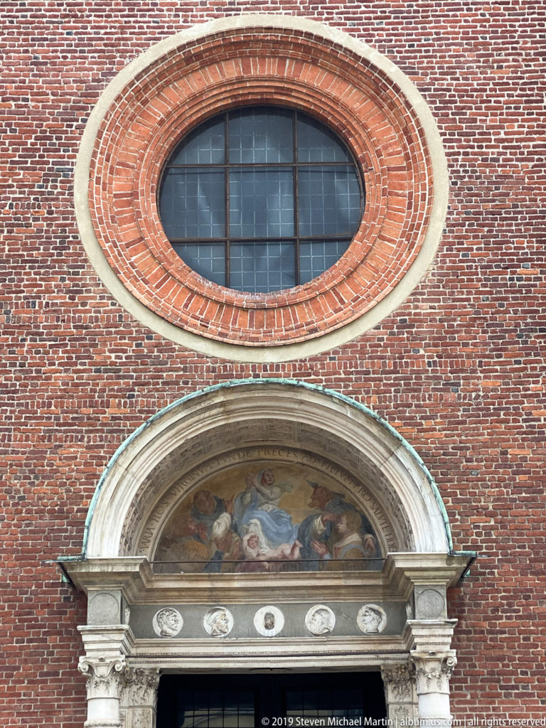 Main portal of Santa Maria delle Grazie with painted semicircular lunette, carved stone surround, and large circular window above.